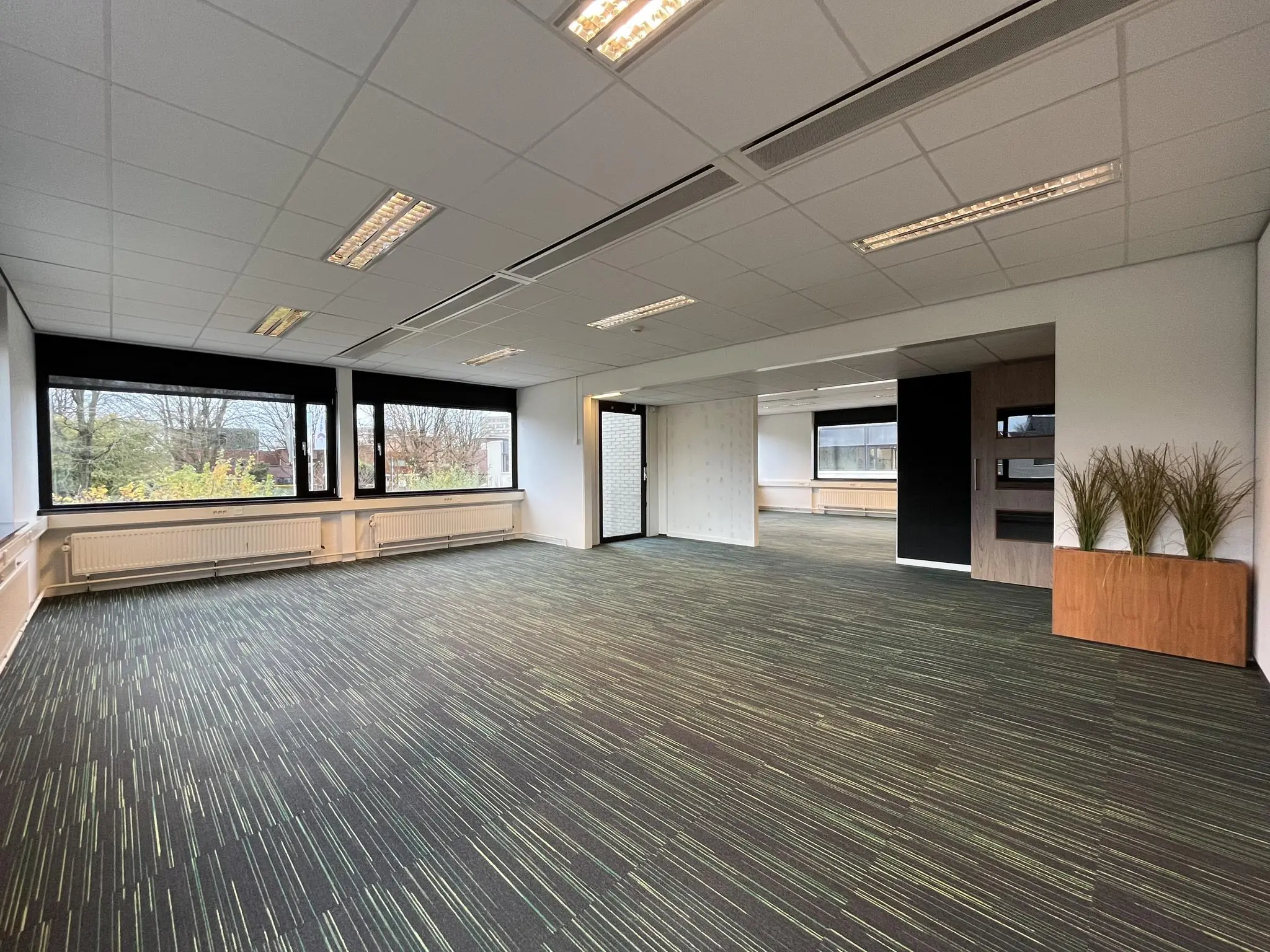 Leeg kantoor met grote ramen, systeemplafond en groen gestreept tapijt aan de Burgemeester Verderlaan.